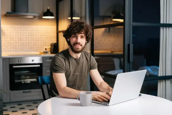 Uśmiechnięty mężczyzna z kręconymi włosami pracuje na laptopie przy stole w kuchni.